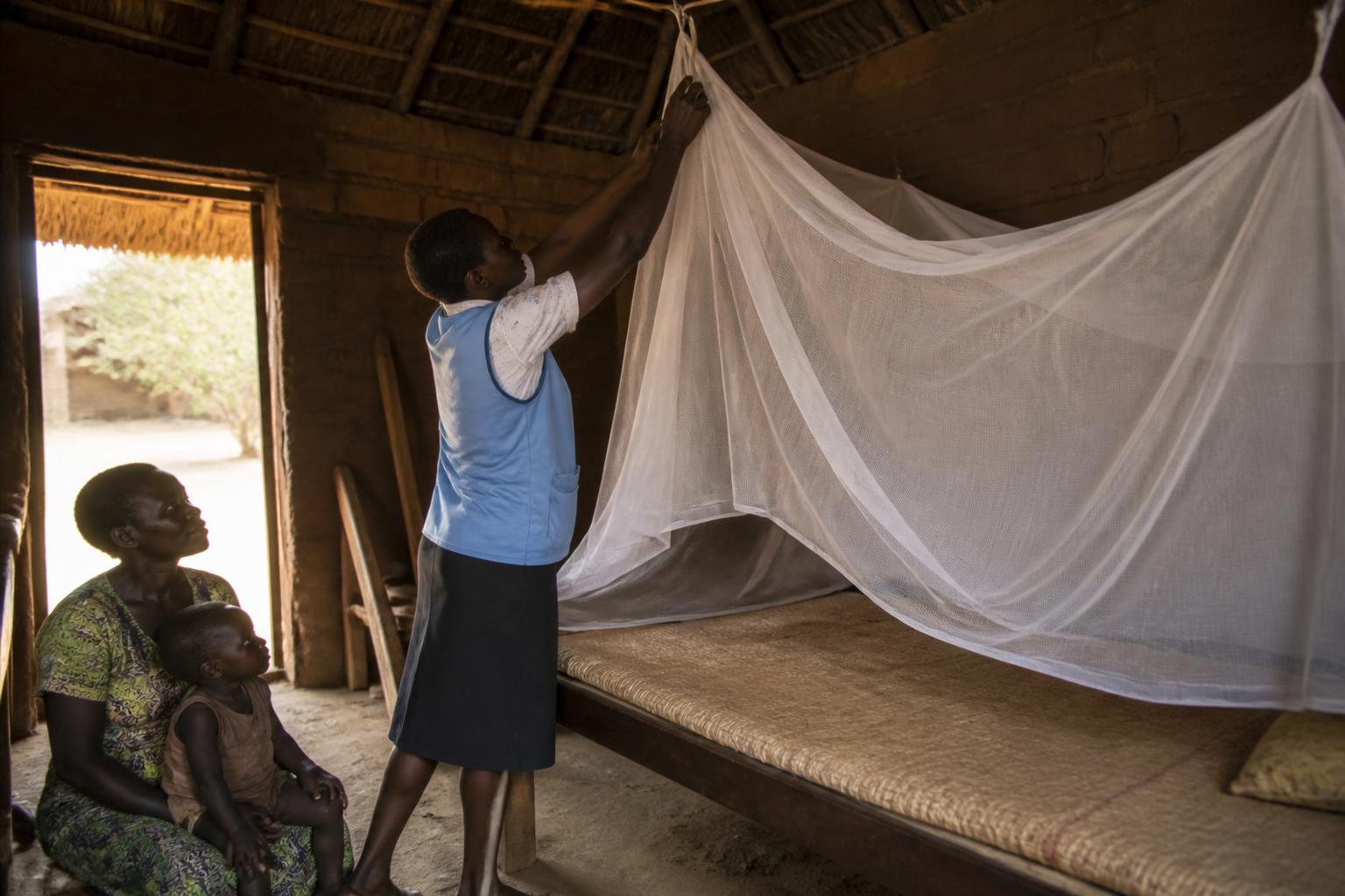 Health worker hanging an insecticide-treated mosquito net over a bed as a mother and child watch